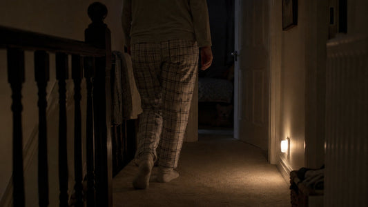A parent in socks walking along a dark upstairs landing at night, lit by a small plug-in night light low on the wall